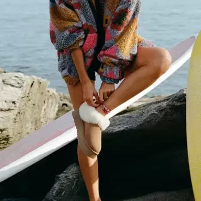 Mulher com casaco fleece colorido e chinelos camel felpudos ao ar livre junto ao mar e pranchas de surf