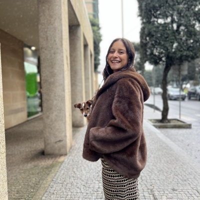 Pessoa com casaco castanho de pelo e cachorrinho numa calçada urbana