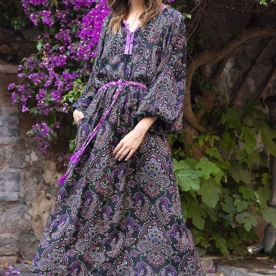 Vestido comprido estampado com fitas roxas e botas pretas em cenário exterior com flores e pedra