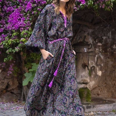 Mulher com vestido longo estampado paisley preto, roxo e verde, cinto roxo e botas pretas em ambiente com flores roxas