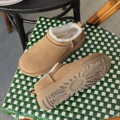 Chinelos de casa castanhos em camurça com interior de pele branca sobre cadeira trançada verde e branca e mesa preta com café e pastelaria