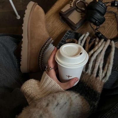 Bota camel de camurça, copo de café, cachecol, auscultadores e carteira sobre fundo de madeira.