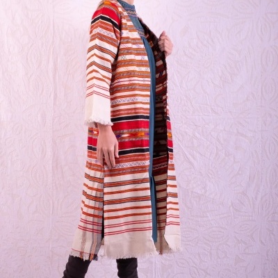 Casaco comprido com riscas coloridas e franjas, usado por mulher com botas pretas, fundo branco texturizado