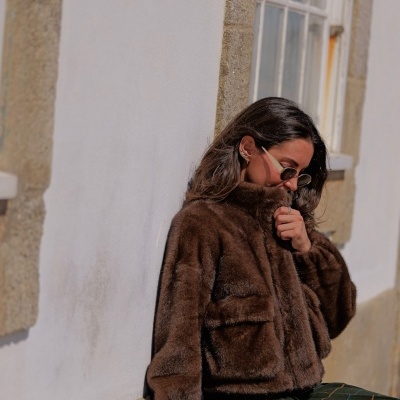 Mulher sentada a usar casaco castanho de pelo sintético e calças quadradas junto a muro branco com janela.