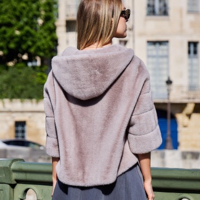 Mulher de costas com casaco de pelo cinza claro e vestido cinza escuro junto a grade verde em área urbana.