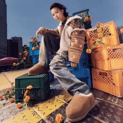 Pessoa com casaco e calças largas vestida com botas castanhas sentada em caixas de plástico coloridas com flores amarelas