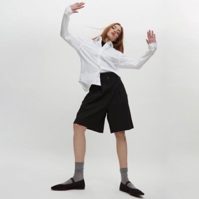 Mulher a usar camisa branca, bermuda preta, meias cinza e sapatos pretos em fundo branco.