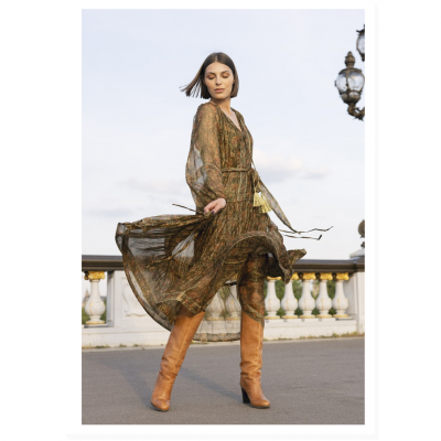 Mulher com vestido longo transparente de padrão abstrato e botas castanhas ao ar livre junto a balaustrada