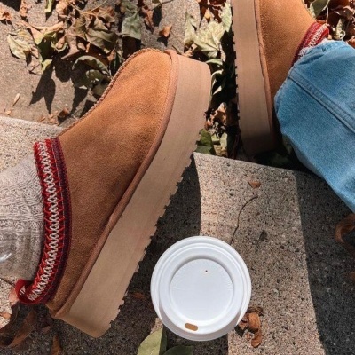 Ténis castanhos slip-on com meias cinza e vermelhas, calças de ganga e copo de café num passeio com folhas secas