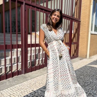 Mulher com vestido maxi branco estampado em padrão geométrico, em calçada de pedra e porta gradeada vermelha ao fundo