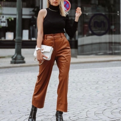 Mulher com top preto de manga única, calças de couro cor caramelo, botas pretas e carteira branca