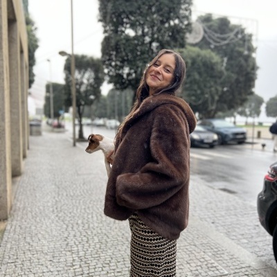 Mulher na rua com casaco castanho felpudo e calças pretas e brancas, segurando cão pequeno.