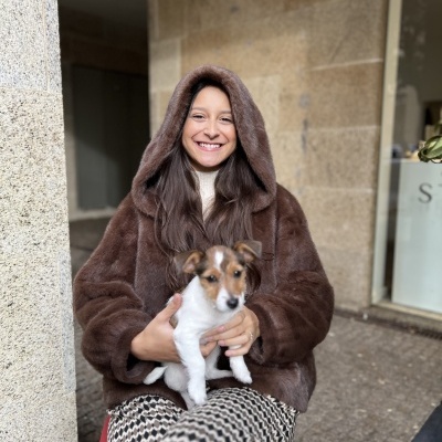 Mulher com casaco castanho felpudo e capuz, calças padrão geométrico e cão pequeno branco e castanho nas mãos
