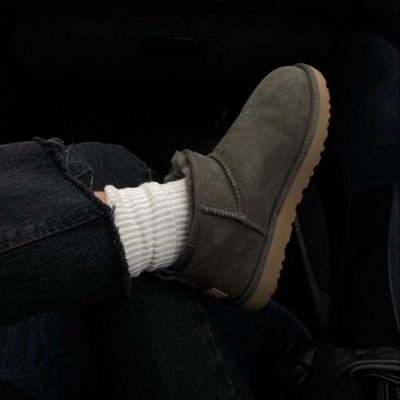 Bota cinza escura de camurça com sola bege clara e interior de pelo, usada com meias brancas e calças de ganga pretas no interior de um carro.