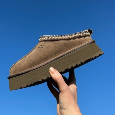 Sapato tipo mule castanho UGG com detalhe em malha e sola de borracha, segurado contra céu azul.