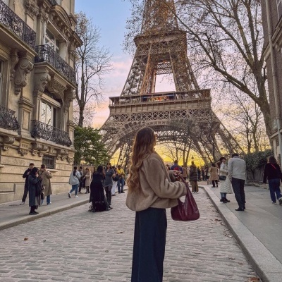 Mulher com casaco castanho e bolsa vermelha na rua com Torre Eiffel ao fundo.