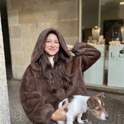 Mulher com casaco peludo castanho e capuz segurando cão pequeno branco com manchas castanhas sentada em ambiente urbano