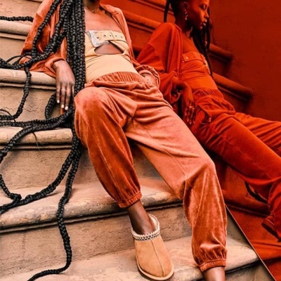 Duas mulheres em roupas de veludo terracota sentadas em escadas, uma usando chinelos castanhos com corrente prateada