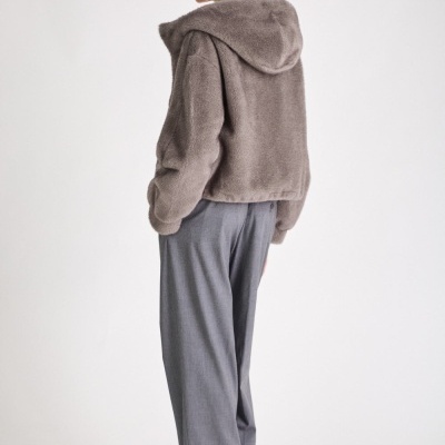 Mulher de costas com casaco de pêlo taupe, calças cinza e sapatos pretos em fundo branco.