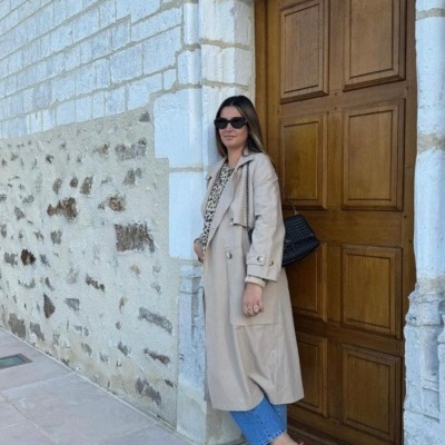 Mulher com casaco bege, camisa animal print, jeans azuis e sapatos castanhos junto a porta de madeira.