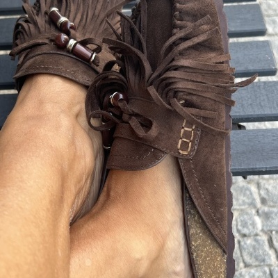 Mocassins castanhos de camurça com franjas e contas, usados por pessoa, em chão de pedras.