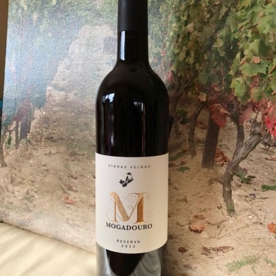 Garrafa de vinho tinto Mogadouro Reserva 2015 com rótulo branco e fundo com imagem de vinhas