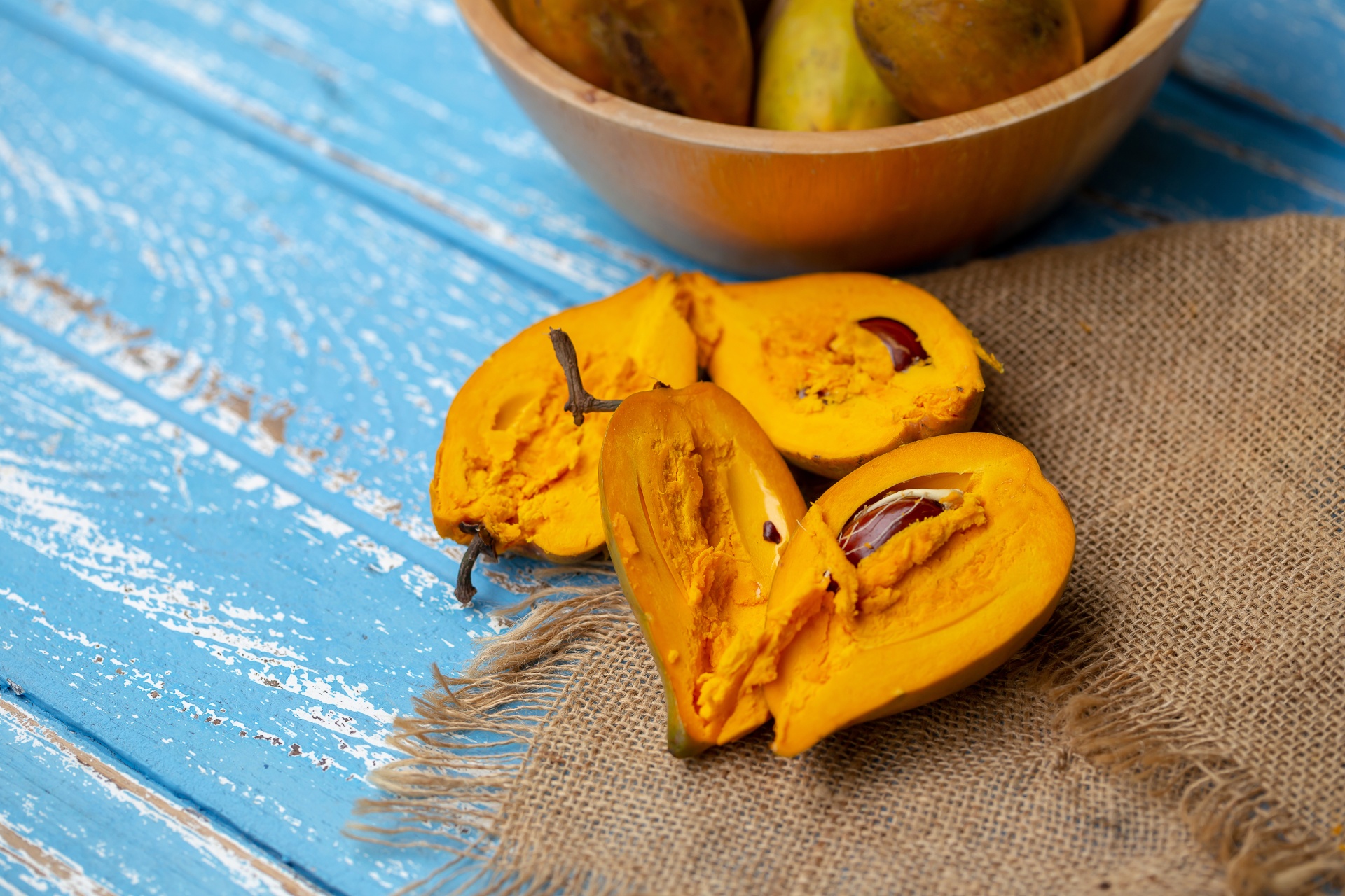 CANISTEL BRUCE ENXERTADO Frutos amarelo-laranja cortados e inteiros sobre pano de juta e madeira azul