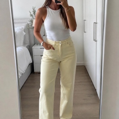 Mulher com top branco, calças amarelas claras e sapatilhas brancas em quarto minimalista.