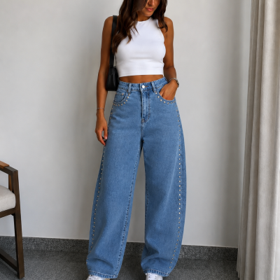 Mulher a usar calças de ganga azuis com tachas, top branco e ténis, em espaço interior com cadeira e cortina