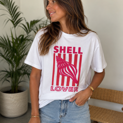 Mulher vestindo t-shirt branca com estampa vermelha SHELL LOVER e concha, com jeans claros, em ambiente interior com planta e banco de madeira.