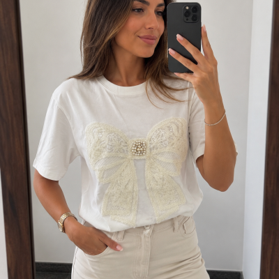 T-shirt branca com laço de renda creme e pérolas, calças bege claras, mulher a tirar selfie em espelho.