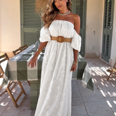 Mulher com vestido longo branco bordado e cinto castanho, em ambiente ao ar livre com mesa e cadeiras de madeira