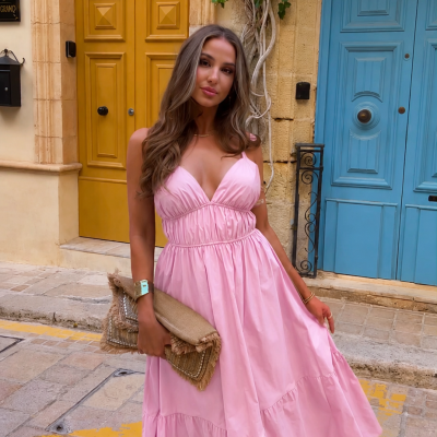 Mulher com vestido longo cor-de-rosa a segurar carteira castanha em rua com portas azul e amarela.