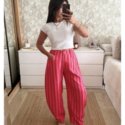 Mulher a experimentar calças largas com riscas rosa e vermelho, t-shirt branca e acessórios, em quarto com cómoda e espelho.