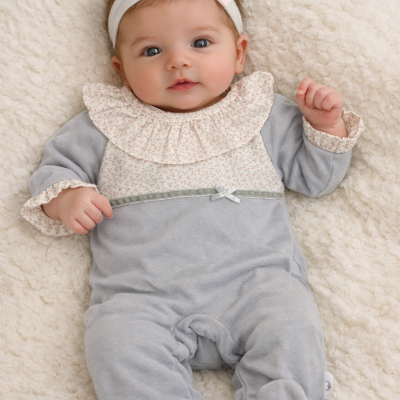 Bebé com macacão cinza detalhado e faixa branca com laço rosa deitada em fundo creme peludo