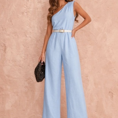 Mulher veste macacão azul claro com um ombro só e cinto branco, segura mala preta e usa sandálias pretas, contra fundo cor de rosa.