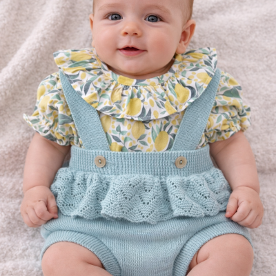 Bebé com roupa estampada com limões e suspensórios azul claro sentado sobre manta.