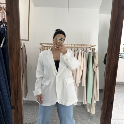 Pessoa a vestir blazer branco, top preto e jeans azul claro em loja com roupas penduradas ao fundo