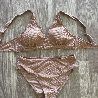 Biquíni bege claro com padrão geométrico texturizado em tecido, composto por top triangular e cuecas, sobre chão de madeira cinzenta.