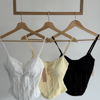 Três tops rendados branco, amarelo claro e preto em cabides de madeira em fundo branco