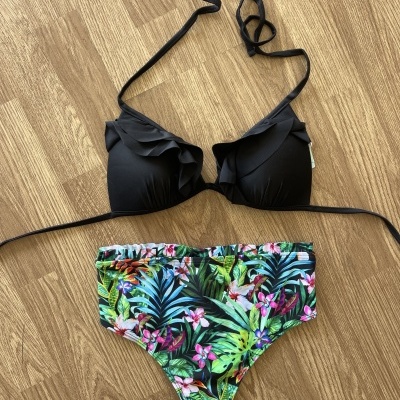 Biquíni preto com parte inferior colorida de padrão tropical sobre chão de madeira.