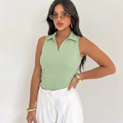 Mulher com blusa verde claro sem mangas e calças brancas posando com acessórios dourados.