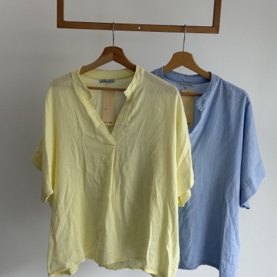 Duas blusas de manga curta, amarelo claro e azul claro, penduradas em cabides de madeira
