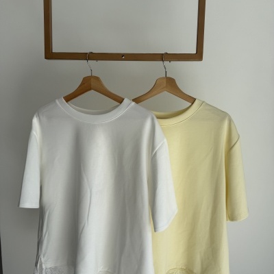 Duas t-shirts em branco e amarelo claro com renda na bainha penduradas em cabides de madeira.