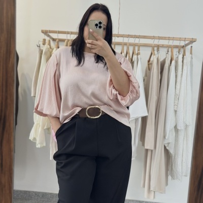 Mulher com blusa rosa de manga comprida e calças pretas a tirar selfie no espelho com cabides e roupa ao fundo.