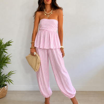 Mulher com conjunto rosa vichy composto por top tomara que caia e calças largas, acessórios dourados, mala bege, planta ao lado