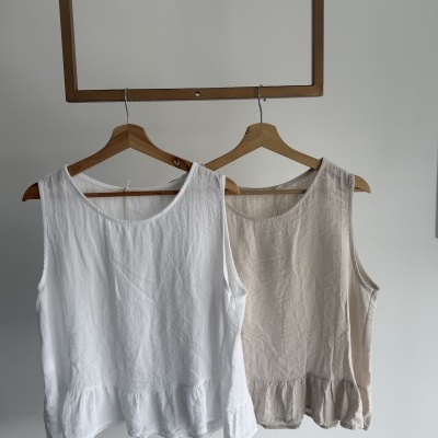 Duas blusas sem mangas, uma branca e outra bege, penduradas em cabides de madeira.