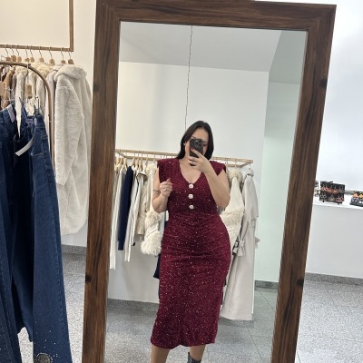 Mulher a usar vestido vermelho com brilho, tirando selfie em espelho de madeira numa loja de roupa com várias peças penduradas e a usar botas pretas com tachas.