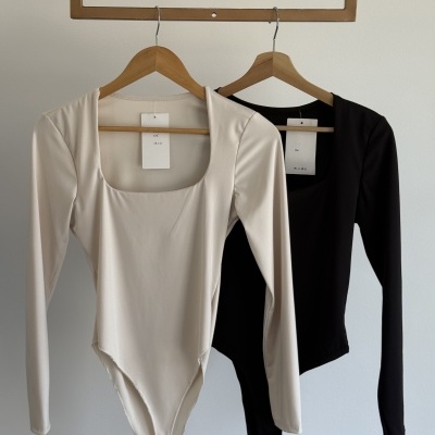 Dois bodys de manga comprida, um creme e outro preto, em cabides de madeira.