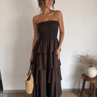 Mulher a usar vestido longo castanho de tecido leve e franzido, segurando mala de palha dentro de casa.